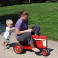 Grown woman riding toy tractor, followed by toddler.
